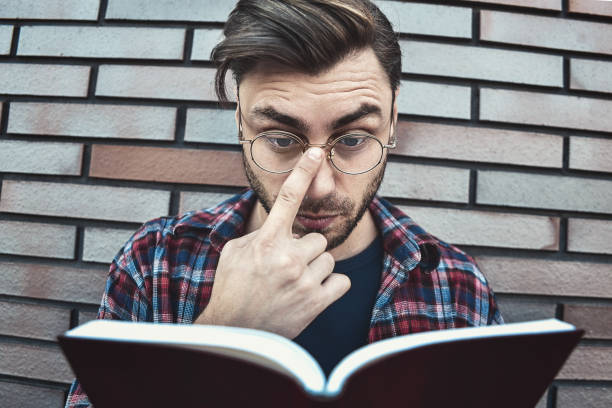 istock Young hipster guy wearing eyeglasses reading a book or note book on brick wall background. 1085616292