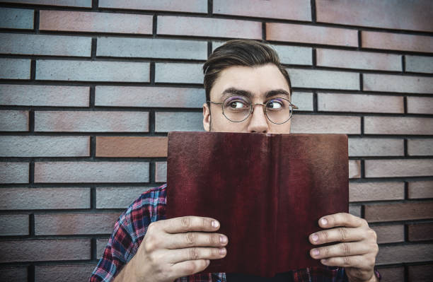 istock Young hipster guy wearing eyeglasses reading a book or note book on brick wall background. 1085616298