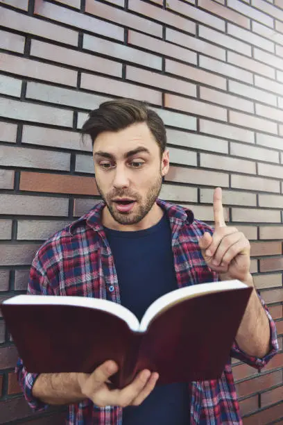 Photo of Young hipster guy reading a book or note book and have some idea on brick wall background.