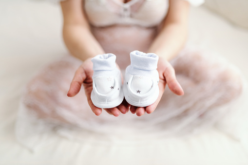 Close up of Caucasian pregnant woman holding baby's footwear in one hand while sitting on the bed with legs crossed.