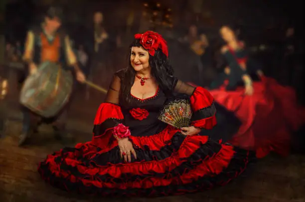 Mature woman dressed in black dress with red flounces and red roses on the hand and head performs gypsy dance pas at the band background. Musicians out of focus.