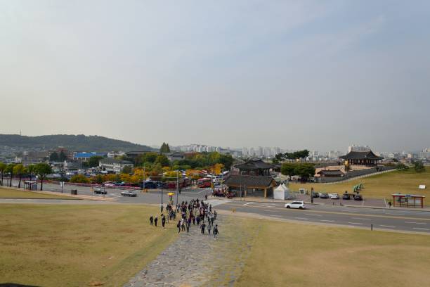 A large mixed group of  teen age children on a field trip to Hwaseong Fortress in Suwon, South Korea, a UNESCO cultural heritage site, advance up the hill to tour the facility.  Meanwhile, a large group of elementary age children in school uniforms wait on the far side of the road to be shown safely across the street to join in the touring activity.  Fortress building and battlements are in the background and the Suwon cityscape is in the far background.