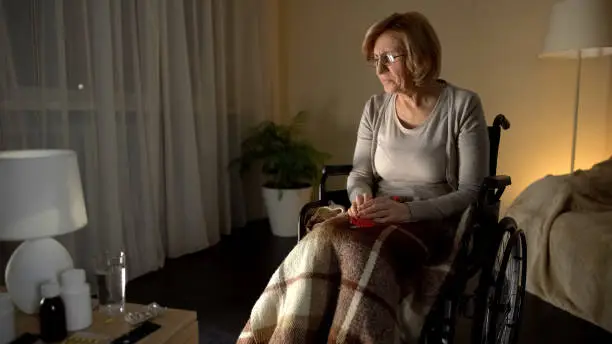 Old tired woman in wheelchair wearing glasses trying to knit, poor eyesight