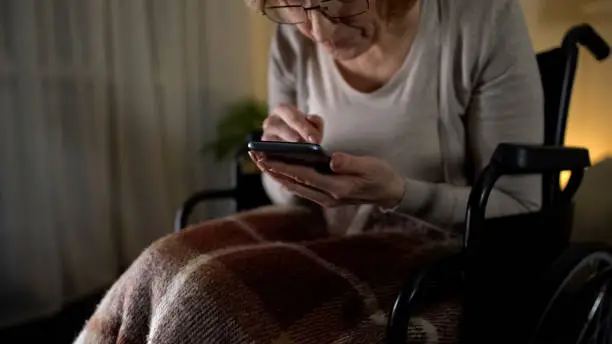 Old woman in wheelchair viewing photos on smartphone, love to grandchildren