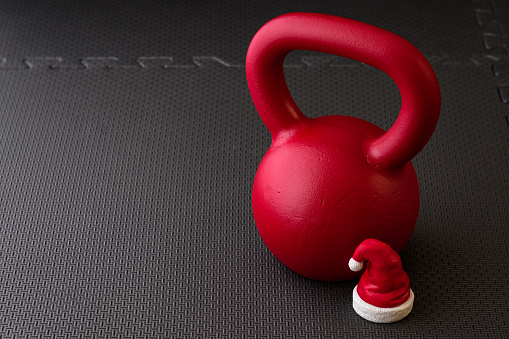 Red kettlebell on a black gym floor with small red and white Santa Claus hat to celebrate Christmas fitness