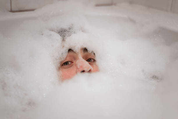 Funny, silly man is completely covered in bubbles in a bath, with only blue eyes and part of his face visible stock photo