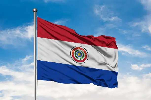 Photo of Flag of Paraguay waving in the wind against white cloudy blue sky. Paraguayan flag.