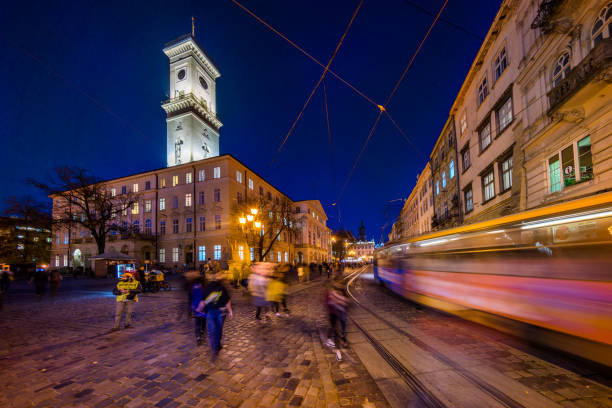 Lviv, Ukraine - October 31, 2018:  view showing people walking around Lviv Old town square, clock tower of city hall, apartment buildings and trams in the tramway can be seen on the area at night