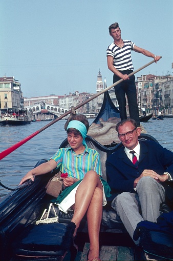 Venice, Veneto, Italy, 1965. A couple (tourists) ride in a Venetian gondola over the Grand Canal of Venice. In the background stands the gondolier.