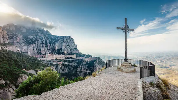 Panorama of christian cross from a Montserrat Mountain Viewpoint in Summer close to Sunset. View to the Monserrat Monastery  in the Mountains. Montserrat is a international well known Monastary near Barcelona, Catalonia, Spain.