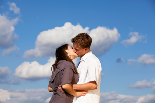 A kissing teenage couple photographed with blue sky and clouds in the background