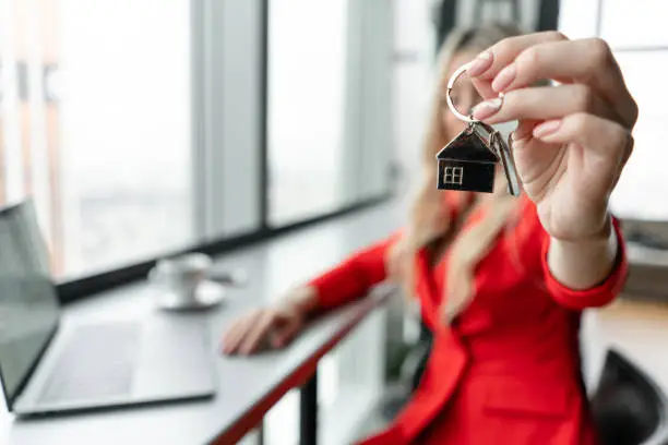 Mortgage concept. Woman in red business suit holding key with house shaped keychain. Modern light lobby interior. Real estate, moving home or renting property
