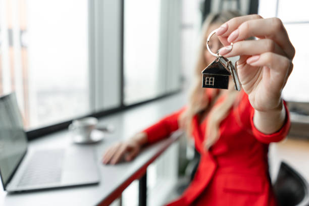 mortgage concept. woman in red coral business suit holding key with house shaped keychain. modern light lobby interior. real estate, hypothec, moving home or renting property. - key real estate key ring house key imagens e fotografias de stock