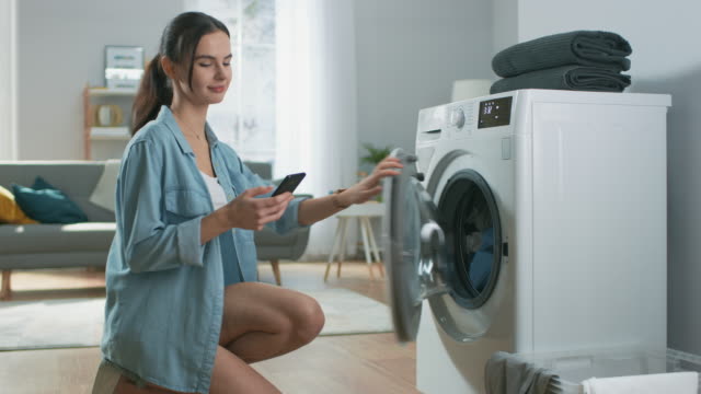 Beautiful Young Woman Sits on Her Knees Next to the Washing Machine. She Loaded the Washer with Dirty Laundry While Using Her Smartphone. Shot in Living Room with Modern Interior.