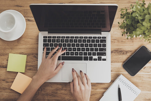 Top view of the working space of a modern business. Female hands are typed on laptop that stands next to cup and notepad on wooden desktop. Digital technologies concept. Creative space.