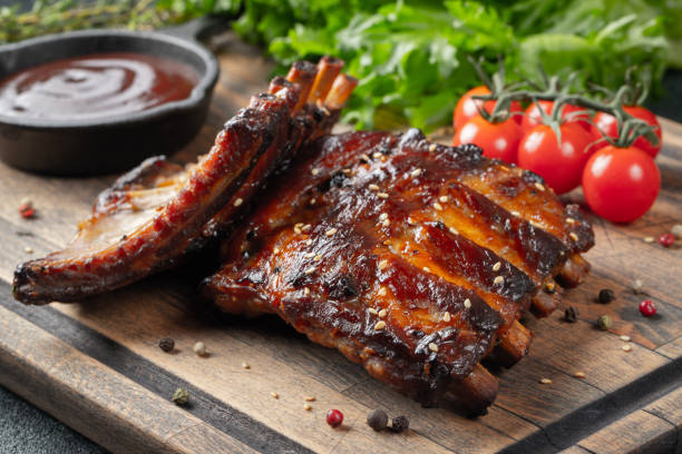 closeup de costillas de cerdo a la parrilla con salsa bbq y caramelizado en miel. sabroso aperitivo a la cerveza en una tabla de madera para presentación en el fondo de hormigón oscuro - costilla fotografías e imágenes de stock