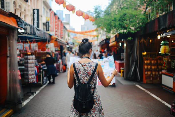 young solo traveler woman in Singapore street market checking the map Rear view image of a young brunette woman. She is enjoying the walk and exploring the city, wearing a casual but fashionable dress, sightseeing and shopping on the Singapore street market. She is holding a large city map, checking out where to go next. travel stock pictures, royalty-free photos & images