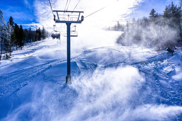 Skiers Riding Chairlift with Snowmaking - Blowing snow on sunny day.