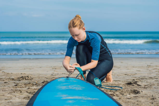 Summer time and active rest concept. Young surfer woman beginner fastens leash across leg, going to surf on big barral waves on ocean, dresseed in boardies, poses aganist sky on hot sunny day. tiempo de verano y concepto de descanso activo. principiante de mujer joven surfista sujeta la correa a través de la pierna, va a practicar surf en olas grandes barral en océano, dresseed en bermudas, plantea contra el cielo en día soleado caliente