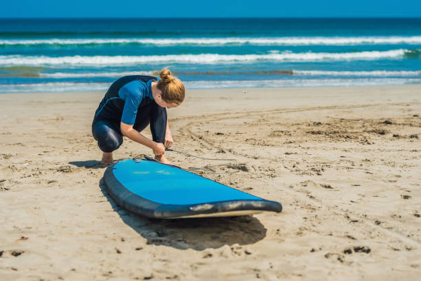 Summer time and active rest concept. Young surfer woman beginner fastens leash across leg, going to surf on big barral waves on ocean, dresseed in boardies, poses aganist sky on hot sunny day. tiempo de verano y concepto de descanso activo. principiante de mujer joven surfista sujeta la correa a través de la pierna, va a practicar surf en olas grandes barral en océano, dresseed en bermudas, plantea contra el cielo en día soleado caliente