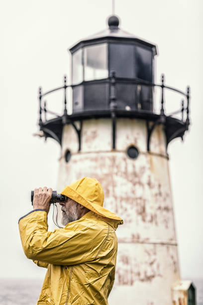 farol keeper vestindo capa impermeável da chuva olhando através de binóculos junto ao farol - looking at view searching looking sea - fotografias e filmes do acervo