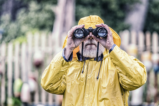 A senior adult man sea captain/lighthouse keeper wearing a yellow rain slicker raincoat and rain hat looking through binoculars at the camera.