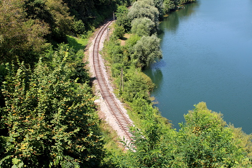 Railway tracks surrounded with tall trees and overgrown vegetation next to clear river