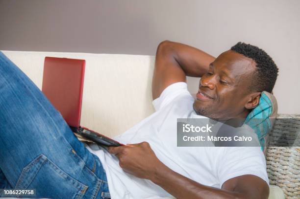 Young Attractive And Happy Successful Black Afro American Man Networking With Laptop Computer At Living Room Couch Smiling Cheerful And Satisfied In Internet Business Success Concept Stock Photo - Download Image Now