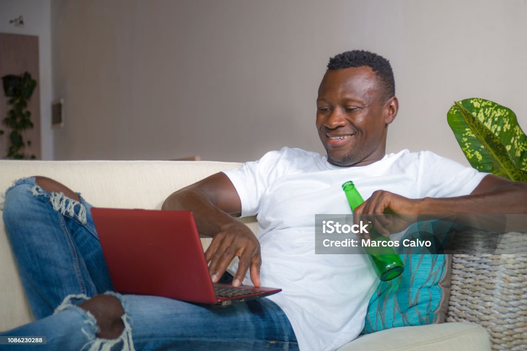 young attractive and happy successful black afro American man networking with laptop computer at living room couch smiling cheerful drinking beer bottle  in internet business success concept 30-39 Years Stock Photo