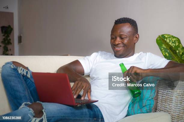 Young Attractive And Happy Successful Black Afro American Man Networking With Laptop Computer At Living Room Couch Smiling Cheerful Drinking Beer Bottle In Internet Business Success Concept Stock Photo - Download Image Now