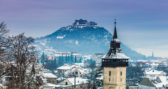 Panoramic color image depicting a snow-covered church spire set amid a snowy, frozen winter landscape and ancient citadel perched on a hill in the town of Deva, in the Transylvania region of Romania. Room for copy space.