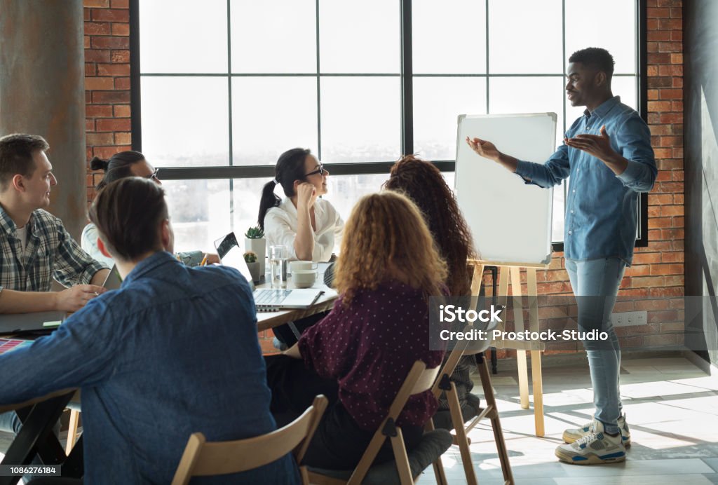 African-american leader lecturing his employees in office African-american team leader is lecturing his employees in loft office using white board, copy space Presentation - Speech Stock Photo