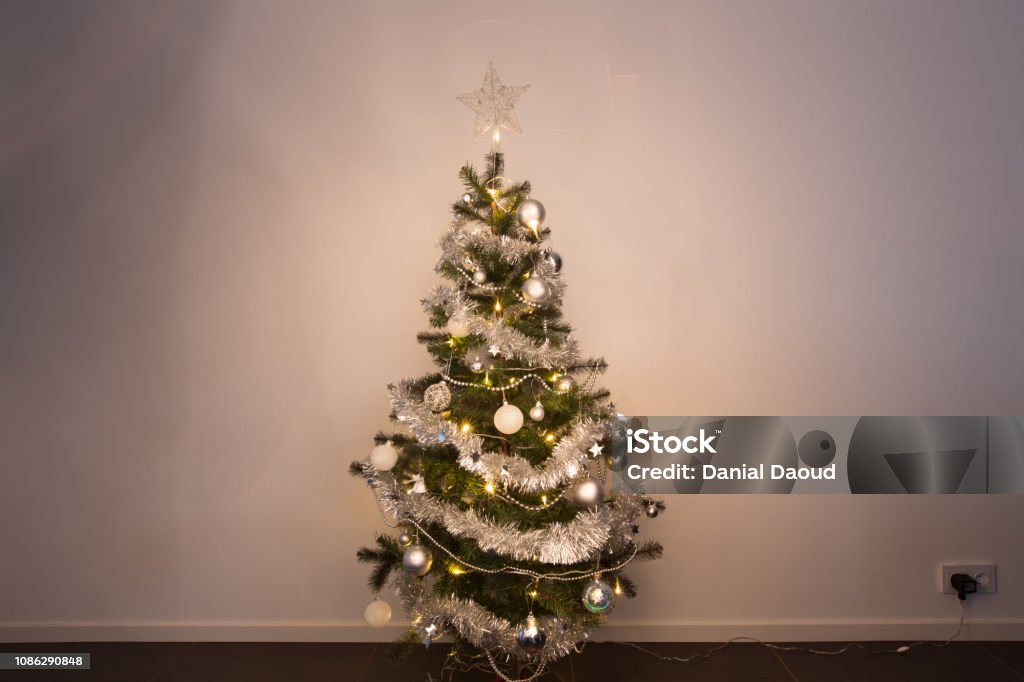 Small Christmas tree standing in front of white wallpaper plugged in and shining warm in middle Christmas Tree Stock Photo