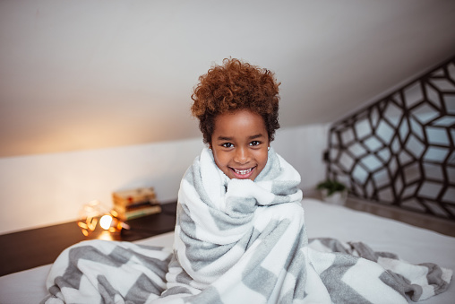 Portrait of a smiling little mixed-race girl with blanket sitting on bed and looking at camera