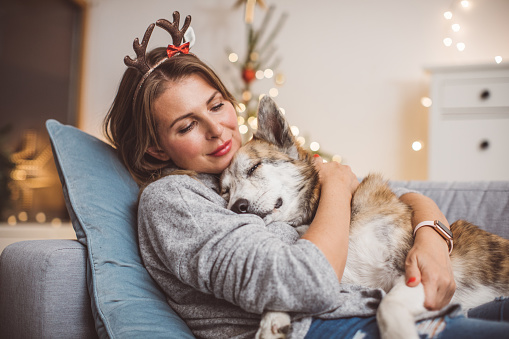 Smiling mature women sitting on couch in living room and wear christmas mask with her dog, christmas tree in background.