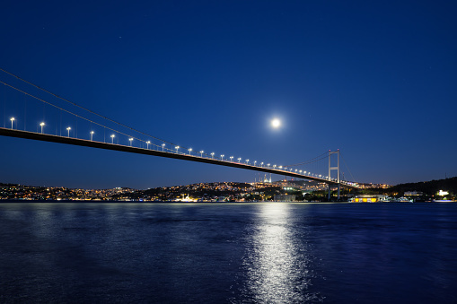 Bosphorus Bridge illuminated by lights and moon at night. Bridge over the Bosphorus to the coast with lighted houses under a bright moon. Istanbul, Turkey.