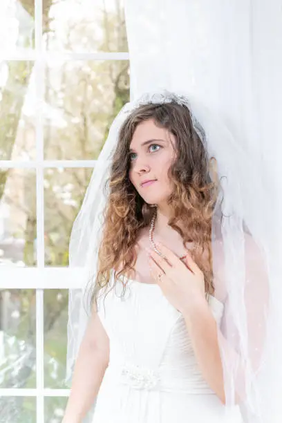 Closeup portrait of young female person, woman, bride in wedding dress, veil, face, pearl necklace, hair, standing, looking up by glass window, white curtains, thinking, engagement ring