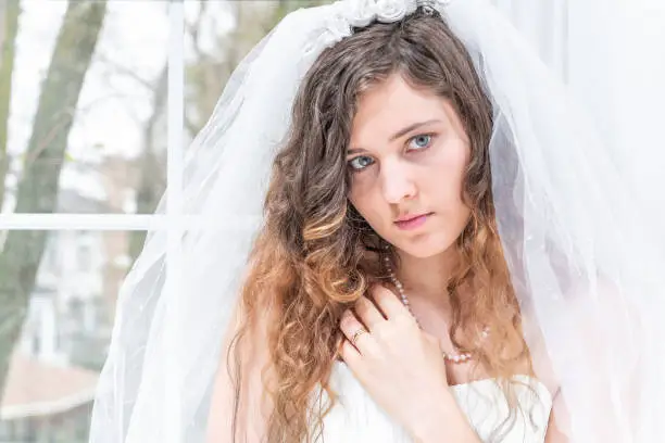 Closeup portrait of young female person, woman, bride in wedding dress, veil, face, pearl necklace, hair, standing, looking up by glass window, white curtains, sad, thinking, engagement ring