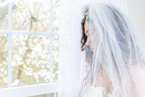 lado de primer plano, retrato de perfil de mujer joven, mujer, novia en vestido de novia, velo, cara, collar de perlas, cabello, de pie, mirando hacia abajo, ve a través de la ventana de cristal, triste, cortinas blancas, pensando - pearl jewelry wedding necklace fotografías e imágenes de stock