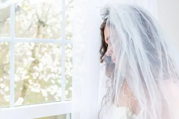 Closeup side, profile portrait of young female person, woman, bride in wedding dress, veil, face, pearl necklace, hair, standing, looking down, view through glass window, sad, white curtains, thinking