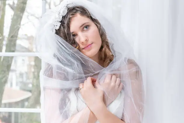 Closeup portrait of young female person, woman, bride in wedding dress, holding, wrapping, covering around shoulders with wrapped veil, by window, white curtains, looking up, in engagement ring