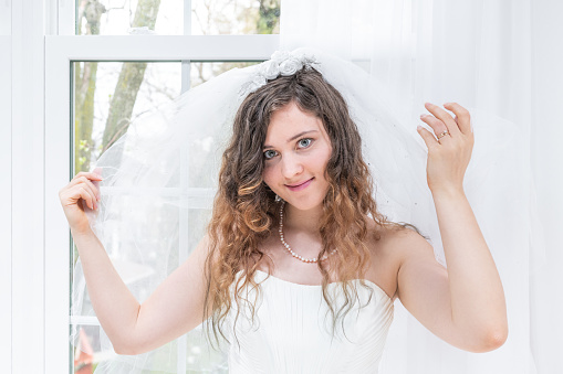 Closeup portrait of young female person, woman, bride in wedding dress, holding veil, hands, face, pearl necklace, standing by window, white curtains, happy, smiling in engagement ring