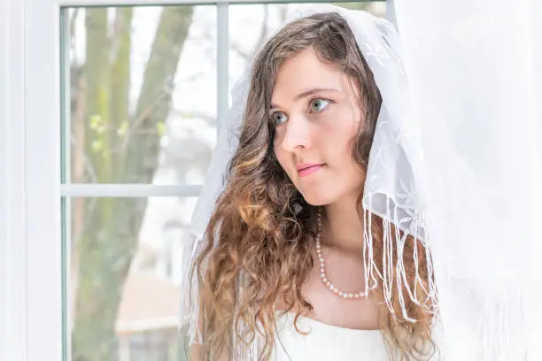 Closeup portrait of young female person, woman, bride in wedding dress, veil, shawl, face, pearl necklace, standing by window, white curtains, looking up, thinking