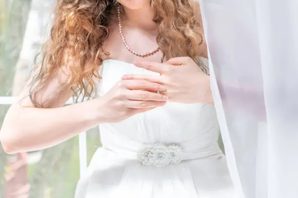 Closeup portrait of young female person, woman, bride in wedding dress, pearl necklace, standing by window, white curtains, taking off engagement diamond ring from finger with hands