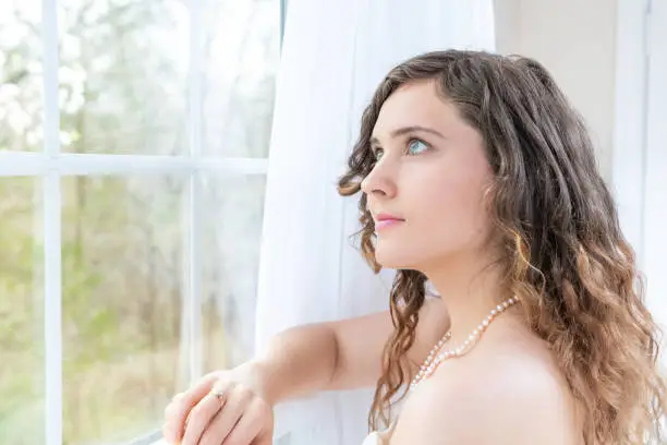 Closeup side, profile portrait of young female person, woman, bride in wedding dress, pearl necklace, hair, face standing, looking up, through glass window, white curtains at view, thinking, leaning