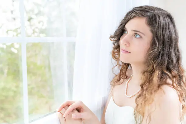 Closeup side, profile portrait of young female person, woman, bride in wedding dress with pearl necklace, hair, face standing, looking through glass window, white curtains at view, thinking