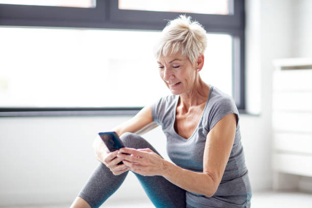 istock Modern short blonde hair mature woman using mobile phone while relax after exercise in corner of room with window at background 1086521388