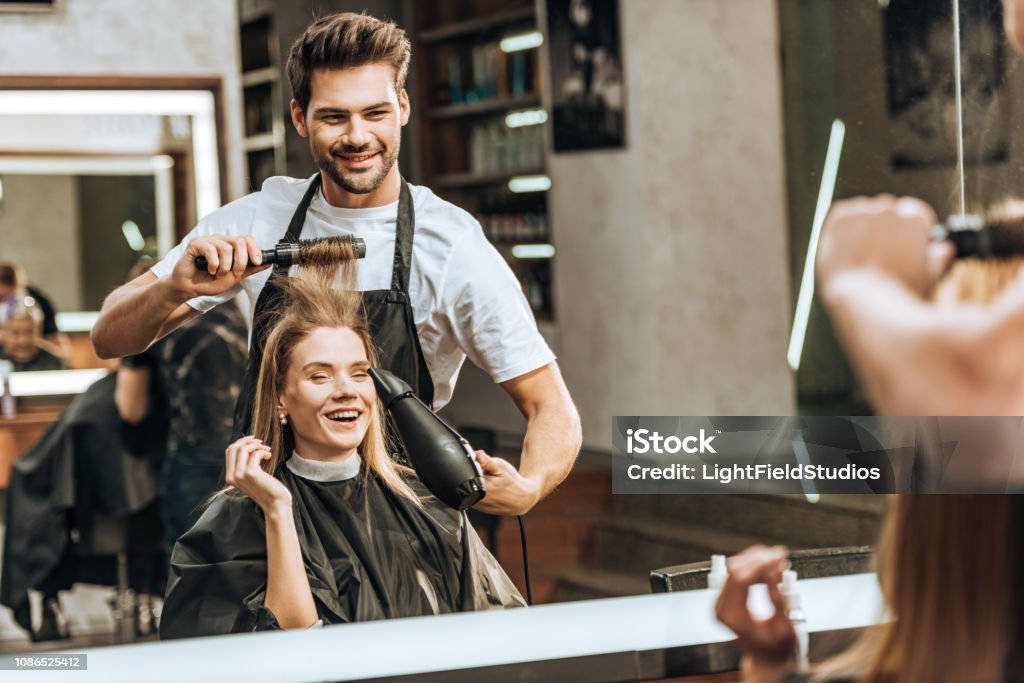 sonriente a joven estilista peinando y secando el pelo a mujer joven feliz en el salón de belleza - Foto de stock de Peluquero libre de derechos