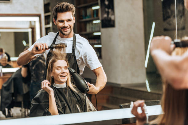 sorridente giovane parrucchiere pettinare e asciugare i capelli alla giovane donna felice nel salone di bellezza - salone di parrucchiere foto e immagini stock