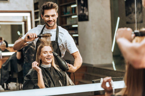 smiling young hairstylist combing and drying hair to happy young woman in beauty salon smiling young hairstylist combing and drying hair to happy young woman in beauty salon hairdresser stock pictures, royalty-free photos & images